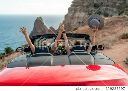 A red convertible car with two people in it, one of whom is waving. The car is driving on a road near the ocean. 119227397