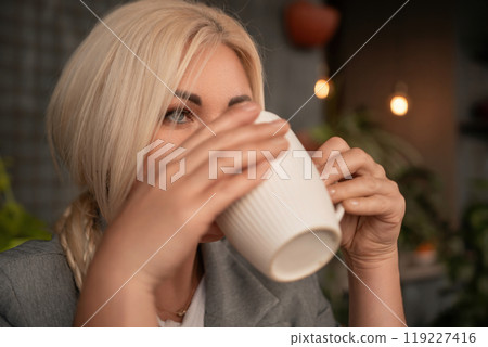 A blonde woman is drinking coffee from a white cup. She is wearing a gray jacket and a necklace. A blonde woman is drinking coffee from a white cup. She is wearing a gray jacket and a necklace. 119227416