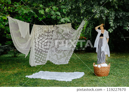 Woman stands in a lush green garden, hanging intricate lace fabric on a clothesline 119227670