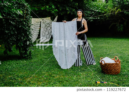 Woman stands in a lush green garden, hanging intricate lace fabric on a clothesline 119227672
