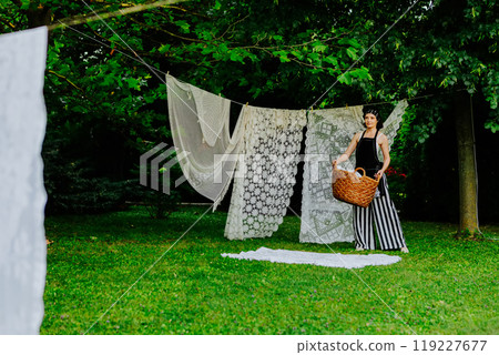 Woman stands in a lush green garden, hanging intricate lace fabric on a clothesline 119227677