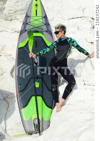 A beautiful sup surfer girl in a wetsuit on the ocean shore against a beautiful blue sky poses sexually with a paddle A beautiful sup surfer girl in a wetsuit on the ocean shore against a beautiful blue sky poses sexually with a paddle 119237242