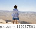 A young woman with short hair stands on a rocky cliff, wearing a light shirt and denim dress, looking out over an expansive desert with distant mountains and clear skies. 119237501