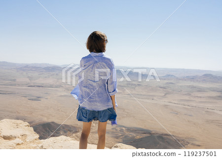 A young woman with short hair stands on a rocky cliff, wearing a light shirt and denim dress, looking out over an expansive desert with distant mountains and clear skies. 119237501