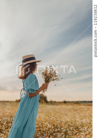Girl in blue linen dress and and canotier straw hat stands back and holds bouquet of flowers in daisy field 119238612