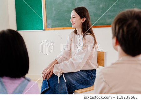 Smiling teacher engages with students in informal classroom circle. Young educator fosters positive learning environment through approachable discussion format in elementary setting Smiling teacher engages with students in informal classroom circle. Young educator fosters positive learning environment through approachable discussion format in elementary setting 119238665