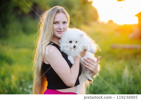 Young blonde woman holds her white fluffy dog in the evening sunlight with a meadow in the background Young blonde woman holds her white fluffy dog in the evening sunlight with a meadow in the background 119238905