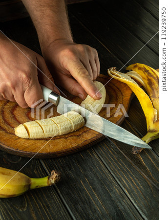 Male hands cutting ripe banana with knife on kitchen board. Concept of healthy breakfast with fruits. Advertising space 119239750