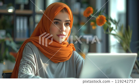 A woman in a hijab working on a laptop in an office. 119242757