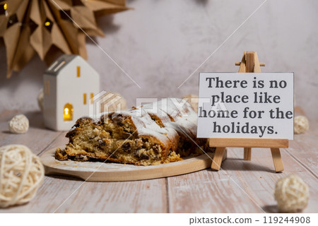 Greeting card with message THERE IS NO PLACE LIKE HOME FOR THE HOLIDAYS.Christmas Stollen in Christmas lights and decoration. Traditional sweet fruit loaf with icing sugar festive pastry dessert 119244908