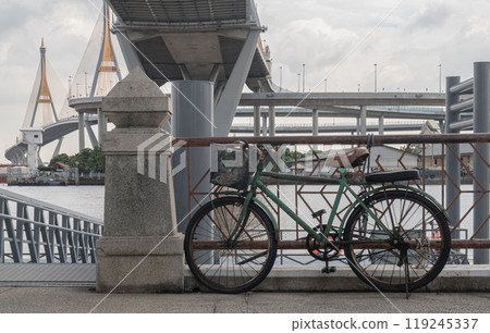 An old bicycle parked on the side in the ferry pier with Bhumibol suspension bridge cross over Chao Phraya River in the background. 119245337