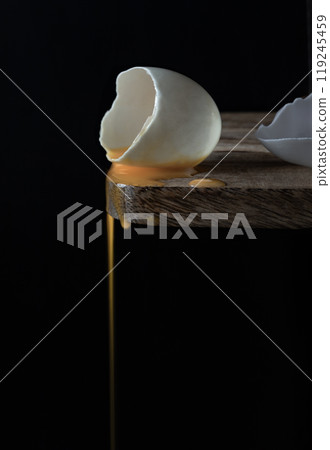 Conceptual image of a broken white eggshell with dripping egg yolk on black background. Conceptual image of a broken white eggshell with dripping egg yolk on black background. 119245459
