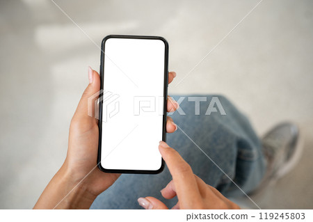 A woman in jeans sitting indoors and using her smartphone, with a white screen mockup displayed. 119245803
