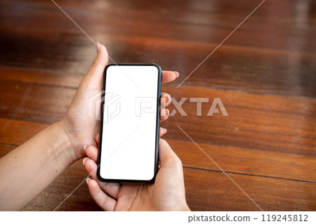 A close-up of a woman sitting at a wooden table indoors and using her smartphone. 119245812