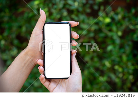 A close-up of a woman's hand holding a smartphone, set against a blurred background of green bushes. 119245819