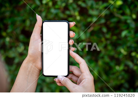 A close-up of a woman's hand holding a smartphone, set against a blurred background of green bushes. A close-up of a woman's hand holding a smartphone, set against a blurred background of green bushes. 119245820