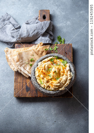 Traditional Jewish hummus served in a stone bowl, garnished with olive oil, paprika, and parsley, alongside flatbreads and fresh herbs on a gray table 119245995