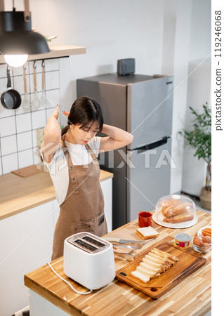 A positive Asian woman is tying her hair while cooking, making toast, and preparing her breakfast. 119246068