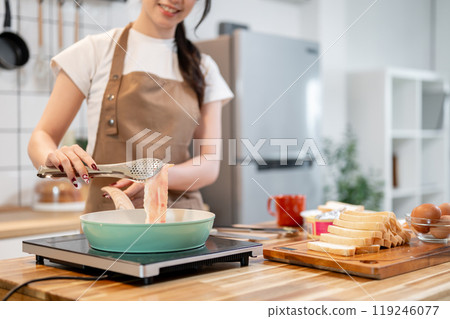 A woman holding tongs, flipping a slice of bacon in a pan while making her breakfast in the kitchen. A woman holding tongs, flipping a slice of bacon in a pan while making her breakfast in the kitchen. 119246077
