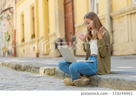 Caucasian young woman using laptop computer celebrating win good message news sitting in city street Caucasian young woman using laptop computer celebrating win good message news sitting in city street 119249901