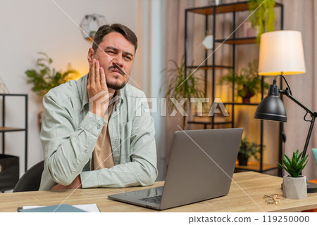 Caucasian young man freelancer sitting at home office table feeling toothache touching mouth at home Caucasian young man freelancer sitting at home office table feeling toothache touching mouth at home 119250000