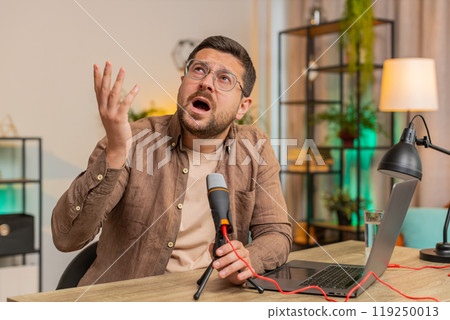 Young positive Caucasian man blogger singer using laptop while singing into microphone at table 119250013