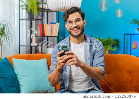 Indian Arabian young man sitting on sofa use smartphone share messages on social media application 119250079