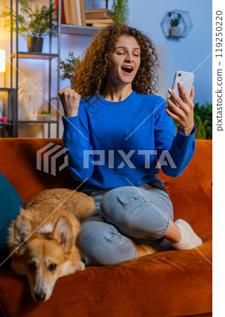 Surprised woman browsing smartphone celebrating success winning lottery sitting beside corgi dog 119250220