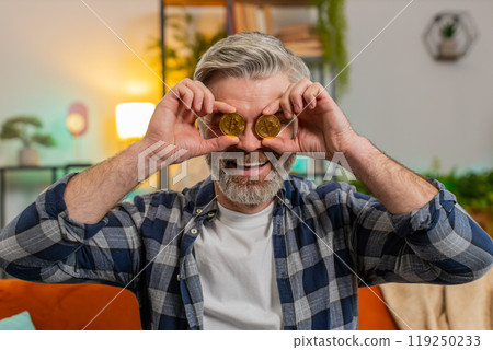 Happy middle-aged man successful stock trader holds covering eyes with golden bitcoins at home room Happy middle-aged man successful stock trader holds covering eyes with golden bitcoins at home room 119250233