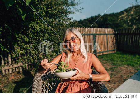 Senior woman is sitting in the garden, eating salad from her own harvest. Older woman is enjoying peaceful lunch alone. Senior woman is sitting in the garden, eating salad from her own harvest. Older woman is enjoying peaceful lunch alone. 119251497