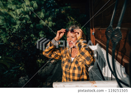 Beautiful older woman is sitting in front of her cabin, enjoying autumn sun rays on her face. 119251505