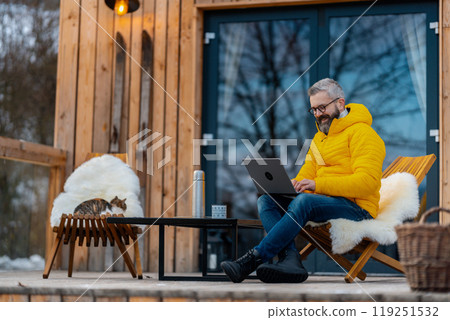 Man working from cozy cabin in mountains, sitting on terrace with laptop, enjoying cup of coffee. Concept of remote work from peaceful location. Hygge at work. 119251532