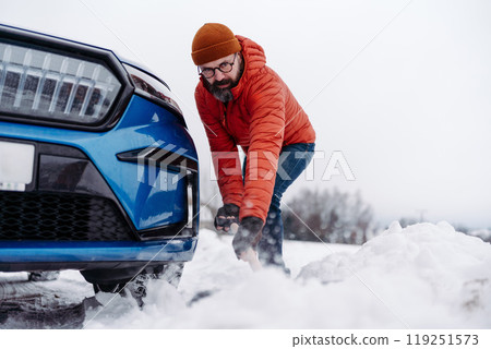 Man freeing his car stuck in the snow with shovel, getting car out from snow drift. Scooping and throwing snow, shoveling. 119251573
