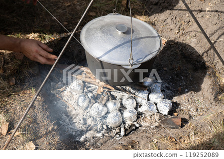 Potatoes wrapped in foil are baked in coals. Camping pot with lid. Cooking food on a campfire on a hike. Fire coals. Potatoes wrapped in foil are baked in coals. Camping pot with lid. Cooking food on a campfire on a hike. Fire coals. 119252089