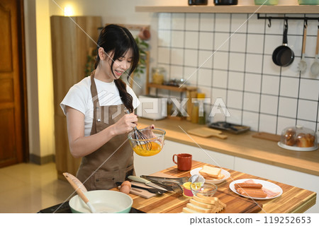 Happy young Asian woman whisking eggs in a glass bowl, preparing breakfast in a kitchen 119252653