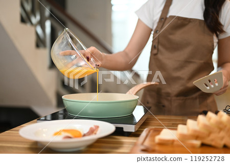 Young woman in apron pouring a beaten eggs from a glass bowl into a frying pan 119252728