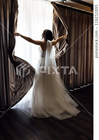 The girl stands with her back near the window and opens the curtains with her hands The girl stands with her back near the window and opens the curtains with her hands 119252895