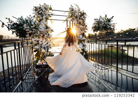 A brunette bride in a white dress holds a bouquet and walks to the wedding arch, among many white chairs. Wedding ceremony in nature. Celebration A brunette bride in a white dress holds a bouquet and walks to the wedding arch, among many white chairs. Wedding ceremony in nature. Celebration 119253342