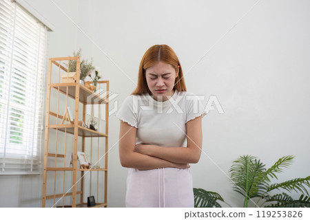 Young Woman Experiencing Stomach Ache at Home, Clutching Her Abdomen in Pain, Standing in a Modern Living Room Young Woman Experiencing Stomach Ache at Home, Clutching Her Abdomen in Pain, Standing in a Modern Living Room 119253826