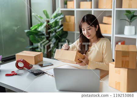 Young Woman Checking Customer Orders for Online Sales in a Modern Home Office with Packaging Supplies 119253868