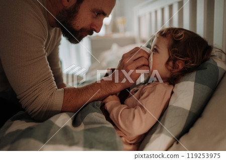 Sick girl with cold lying in the bed. Father helping little daughter to blow her nose. Sick girl with cold lying in the bed. Father helping little daughter to blow her nose. 119253975