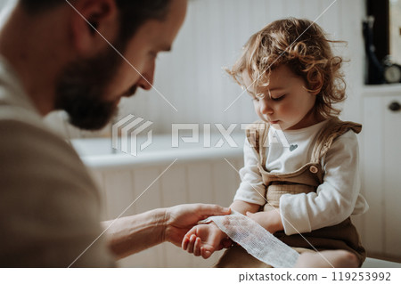 Father is bandaging his daughter's injured wrist in bathroom, concentrating while using a bandage. 119253992