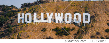 The Hollywood Sign stands prominently on a dry, grassy hillside with sparse vegetation and trees, symbolizing the entertainment industry in Los Angeles. 119254045