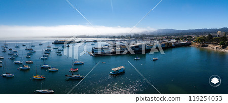 An aerial view captures Monterey, California's harbor filled with sailboats and yachts. Waterfront buildings line the shore, with fog over distant hills. An aerial view captures Monterey, California's harbor filled with sailboats and yachts. Waterfront buildings line the shore, with fog over distant hills. 119254053
