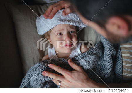 Father taking care of sick son with fever and cold, holdig cold cloth to his forehead. Boy lying on sofa, feeling tired. Top view. Father taking care of sick son with fever and cold, holdig cold cloth to his forehead. Boy lying on sofa, feeling tired. Top view. 119254054