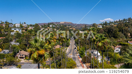 The image captures the Hollywood district in Los Angeles, featuring the Hollywood Hills and Sign. A tree lined street with palm trees and houses is visible. 119254079