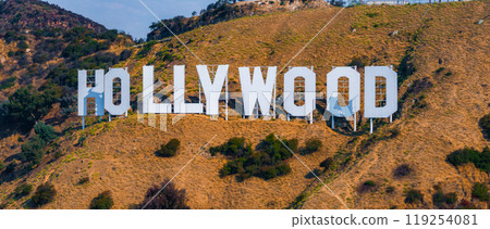 The Hollywood Sign is prominently displayed on the dry, grassy hills of Mount Lee, surrounded by green shrubs and trees under a clear blue sky. The Hollywood Sign is prominently displayed on the dry, grassy hills of Mount Lee, surrounded by green shrubs and trees under a clear blue sky. 119254081