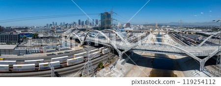 The image captures the Los Angeles River's concrete channel with the Sixth Street Viaduct. Downtown skyscrapers and freight trains add to the urban scene. 119254112