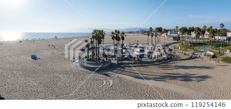 Venice Beach in Los Angeles features a bustling skate park, surrounded by palm trees and visitors. The sandy beach extends to the ocean, with distant mountains visible. Venice Beach in Los Angeles features a bustling skate park, surrounded by palm trees and visitors. The sandy beach extends to the ocean, with distant mountains visible. 119254146