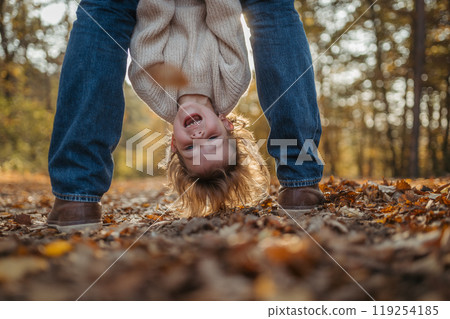 Father holding little girl by legs heand down, playing during walk in autumn forest. Dad and girl having fun outdoors. 119254185